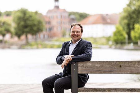 Der Geschäftsführer der Eutin Tourismus GmbH, Jan-Philipp Willers, sitzt auf einer Bank, im Hintergrund sind der See und das Schloss zu sehen