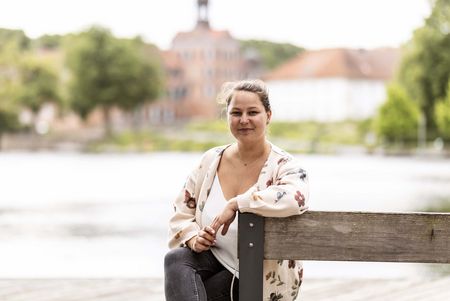Die Mitarbeiterin der Tourist-Info Eutin Janett Paulauskas sitzt auf einer Bank, im Hintergrund sind der See und das Schloss zu sehen