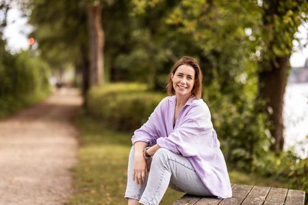Frau mit lila Hemd sitzt auf Picknickbank vor Bäumen