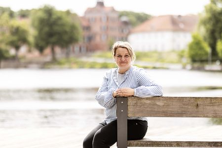 Die Mitarbeiterin der Tourist-Info Eutin Andrea Bahr sitzt auf einer Bank, im Hintergrund sind der See und das Schloss zu sehen